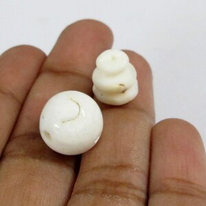 May include: Two off-white, carved beads. One is a sphere with a curved line, and the other is a stacked, cylindrical shape. The beads are held in a hand against a white background. These beads could be used for jewelry making.