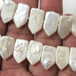 May include: Close-up of a strand of luminous, shield-shaped, off-white pearls. The pearls are strung together, showcasing their irregular, textured surfaces and natural iridescence. The pearls are a creamy white color.