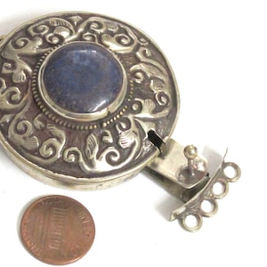 May include: A silver-toned, round pendant with an ornate, engraved design and a central, dark blue stone. The pendant has a clasp and is shown next to a penny for size comparison.