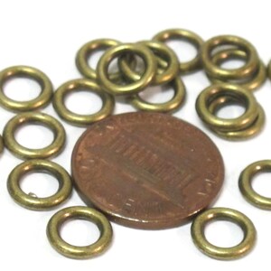 May include: A collection of small, brass-colored metal rings scattered around a penny. The rings are circular with a small hole in the center. The penny is a copper color and shows the profile of a person.