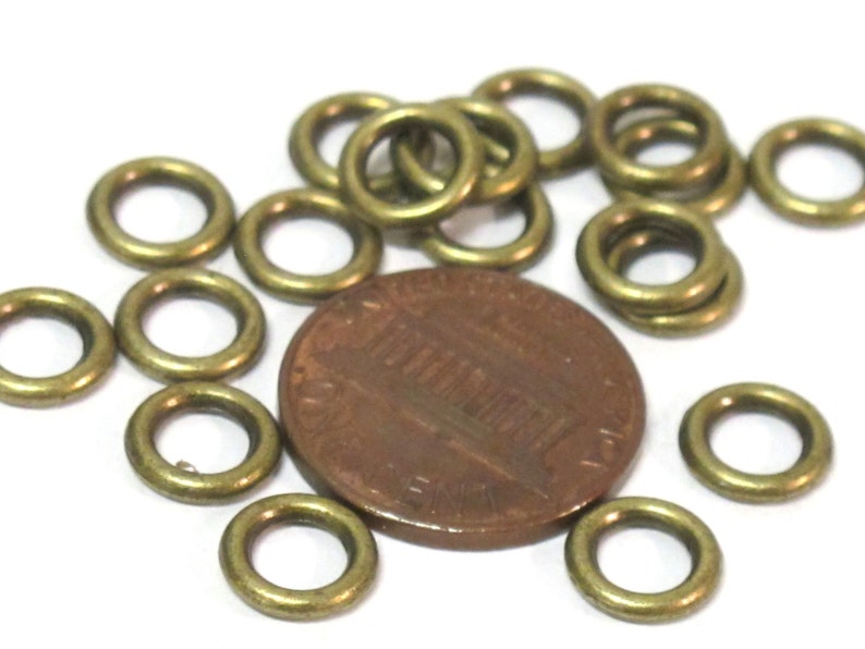 May include: A collection of small, brass-colored metal rings scattered around a penny. The rings are circular with a small hole in the center. The penny is a copper color and shows the profile of a person.