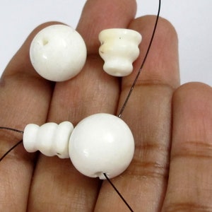 May include: Close-up of white beads and decorative elements. The image shows a variety of shapes, including spheres and a small, cup-like design. The beads are strung on a thin, black thread, suggesting they are for jewelry making or crafts.