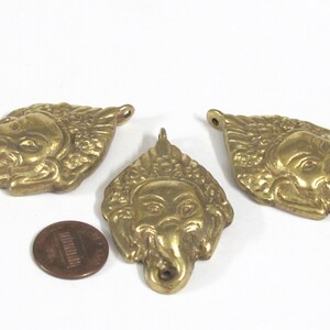 Puede incluir: Tres colgantes de lat&oacute;n con forma de cabeza de elefante, cada uno con un lazo para colgar. Los colgantes presentan rasgos faciales detallados y elementos decorativos. Una moneda est&aacute; al lado de los colgantes para la comparaci&oacute;n de tama&ntilde;o.