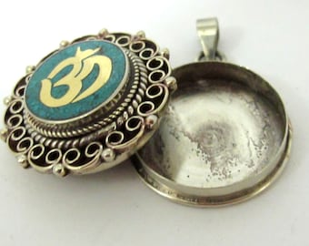 May include: A silver-colored pendant with an open locket. The pendant's front features a turquoise-colored circle with a gold-colored Om symbol. The pendant has an ornate, detailed design around the turquoise circle.