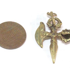 May include: A detailed, gold-colored pendant with an intricate design, featuring a cross-like structure with a blade, a curved element, and a pointed tip. A penny is next to the pendant for size comparison.