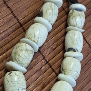 May include: A handcrafted necklace featuring large, off-white, barrel-shaped beads with natural markings, separated by smaller, flat, white disc-shaped beads. The necklace is strung on a black cord, creating a striking contrast. The beads have a unique, organic appearance.