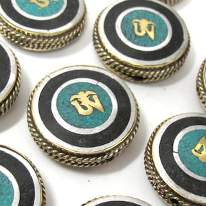May include: Close-up of several round, decorative pendants. Each pendant features a turquoise center with a gold symbol, surrounded by a black band, a silver band, and a gold rope-like edge. The pendants are on a white background.