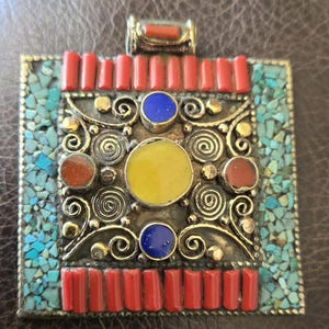 May include: A rectangular pendant with a silver-colored metal frame. The pendant features red, yellow, and blue stones, along with turquoise chips. The design includes spiral and rectangular elements, creating a detailed and colorful accessory.