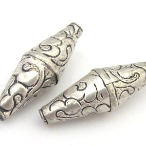 May include: Two silver-colored, elongated beads with intricate, swirling patterns. The beads have a slightly tapered shape and are connected by a band. The design features a detailed, embossed floral or leaf-like motif.
