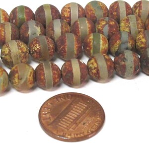 May include: A collection of round, patterned beads in shades of brown, tan, and green, with a lighter stripe encircling each bead. A copper penny is included for size reference. These beads are suitable for jewelry making.