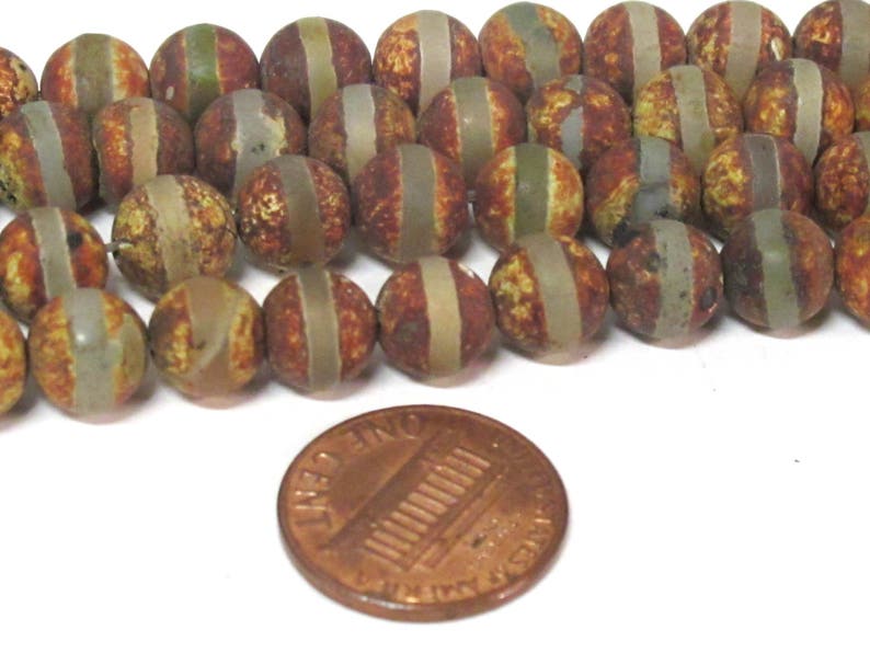 May include: A collection of round, patterned beads in shades of brown, tan, and green, with a lighter stripe encircling each bead. A copper penny is included for size reference. These beads are suitable for jewelry making.
