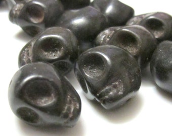 May include: A collection of dark gray skull-shaped beads. Each bead has eye and nose sockets, and a rounded top. The beads are clustered together, with some in sharp focus and others slightly blurred, against a white background.