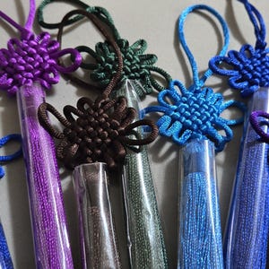 May include: A collection of colorful Chinese knot tassels in shades of blue, purple, green, and brown. Each tassel features a decorative knot and a cylindrical, clear plastic tube containing the tassel threads. The tassels are arranged closely together.