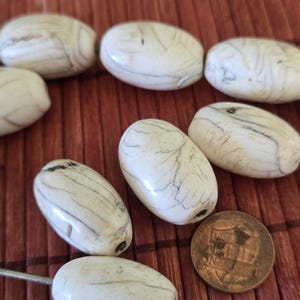 May include: Oval-shaped white beads with gray veining, strung on a silver wire. A copper penny is next to the beads for size comparison. The beads are on a reddish-brown surface.
