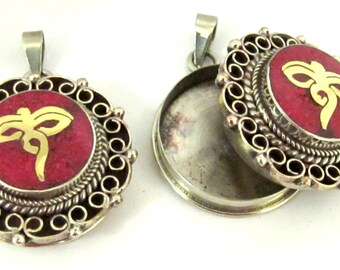 May include: Two silver-tone pendants with a round, red center and a gold-colored symbol. The pendants have an ornate, decorative border. One pendant is open, revealing a hollow interior.