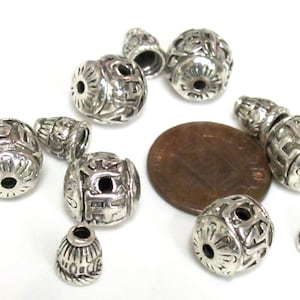 May include: A collection of silver-colored metal beads in various shapes and sizes. The beads have intricate, carved designs and are displayed on a white surface. Some beads are cylindrical, while others are round or cone-shaped.