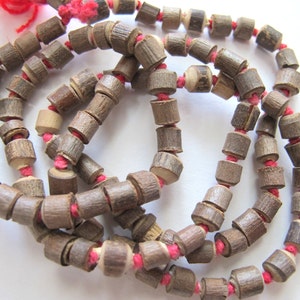 May include: A necklace composed of cylindrical wooden beads strung on a red cord. The beads are a mix of light and dark brown, with a natural wood grain appearance. The red cord is visible between the beads, and a red tassel is at the end.