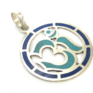 May include: Silver-tone pendant with an Om symbol. The pendant features a circular design with a blue border and a turquoise Om symbol in the center. The pendant has a loop for a chain.