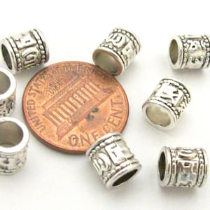 May include: A collection of silver-colored metal beads with etched designs. The cylindrical beads are shown with a penny for size comparison. The beads have a hole running through the center and are decorated with patterns and symbols.