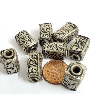 May include: A collection of silver-toned, rectangular beads with intricate black swirl designs. Each bead has a hole running through its center. A penny is included for size reference. These beads are suitable for jewelry making.