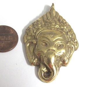 Puede incluir: Un colgante de lat&oacute;n dorado con la forma de una deidad con cabeza de elefante, probablemente Ganesha, con detalles intrincados. El colgante se muestra junto a una moneda de un centavo estadounidense para comparar el tama&ntilde;o.