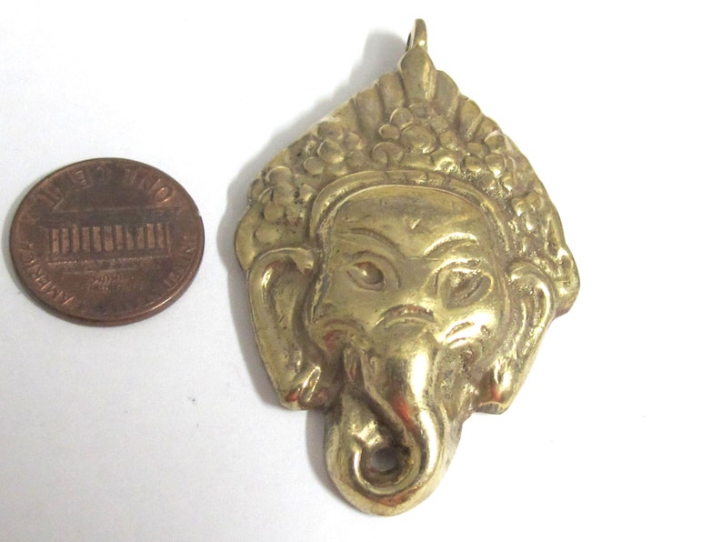 Puede incluir: Un colgante de lat&oacute;n dorado con la forma de una deidad con cabeza de elefante, probablemente Ganesha, con detalles intrincados. El colgante se muestra junto a una moneda de un centavo estadounidense para comparar el tama&ntilde;o.