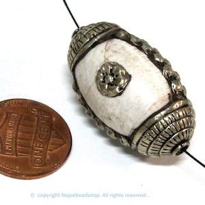 May include: An oval-shaped white bead with silver-colored metal accents. The bead has a floral design and is next to a penny. The bead is strung on a black string. The bead is approximately 1 inch long.