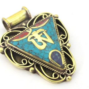 May include: A detailed pendant with a turquoise stone center, featuring a gold Om symbol. The pendant has a gold-colored frame with intricate filigree and red and blue accents. The bail is gold-colored.