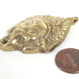 Puede incluir: Colgante de lat&oacute;n con forma de cabeza de elefante con detalles intrincados, incluyendo una trompa y colmillos. El colgante est&aacute; junto a una moneda para comparar el tama&ntilde;o. El colgante mide aproximadamente 5 cm de largo.