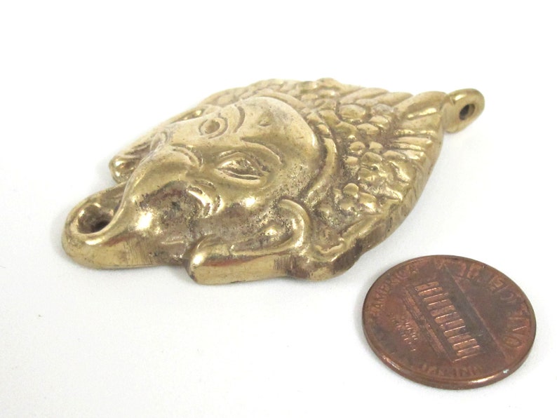Puede incluir: Colgante de lat&oacute;n con forma de cabeza de elefante con detalles intrincados, incluyendo una trompa y colmillos. El colgante est&aacute; junto a una moneda para comparar el tama&ntilde;o. El colgante mide aproximadamente 5 cm de largo.
