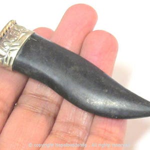 May include: A dark gray pendant with a silver-colored cap and a gold-colored rope design. The pendant is held in a hand against a white background. The pendant has a smooth, polished surface.