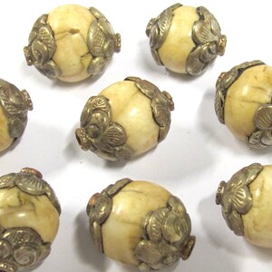 May include: Eight cream-colored, oval-shaped beads with silver-colored floral metal accents. The beads have a slightly textured surface and are arranged against a white background. These beads could be used for jewelry making or other craft projects.