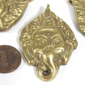 Puede incluir: Primer plano de varios colgantes de lat&oacute;n con la cara de Ganesha, la deidad hind&uacute; con cabeza de elefante. Los colgantes est&aacute;n intrincadamente detallados con un lazo en la parte superior para colgar. Se incluye una moneda como referencia de tama&ntilde;o.