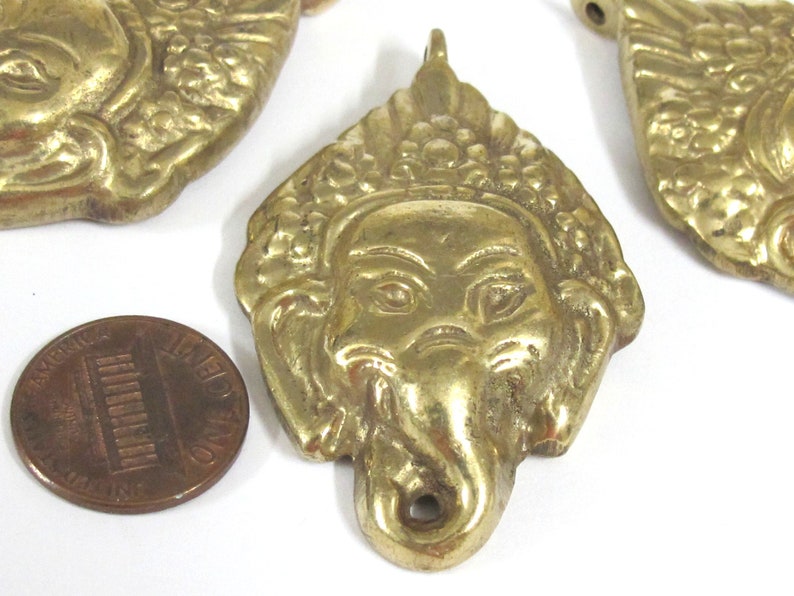 Puede incluir: Primer plano de varios colgantes de lat&oacute;n con la cara de Ganesha, la deidad hind&uacute; con cabeza de elefante. Los colgantes est&aacute;n intrincadamente detallados con un lazo en la parte superior para colgar. Se incluye una moneda como referencia de tama&ntilde;o.