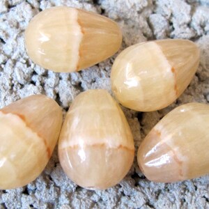 May include: Five polished, teardrop-shaped stones. The stones are a creamy beige color with a lighter, translucent stripe down the center and a hint of orange. The stones are smooth and glossy, suggesting they are gemstones or decorative items.