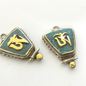 May include: Two triangular pendants with a turquoise inlay and a gold-colored Om symbol. The pendants have a silver-colored frame and a small loop at the top. A small gold-colored bead is at the bottom.