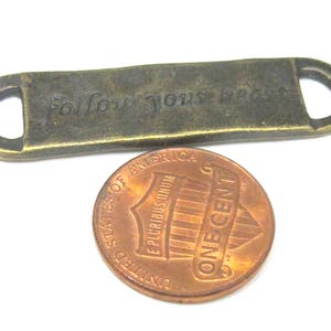 May include: A small, rectangular, bronze-colored metal tag with the engraved phrase "follow your heart". The tag has rounded ends with holes. A copper one-cent coin is next to the tag for size comparison.