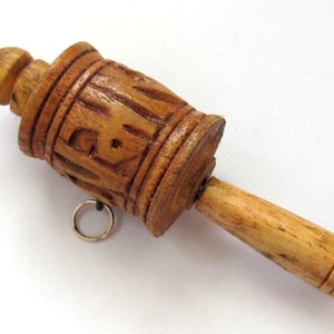 May include: A handcrafted wooden prayer wheel with carved details and a small metal ring. The prayer wheel is light brown and has a cylindrical body with a wooden handle. The top of the wheel has a conical shape.