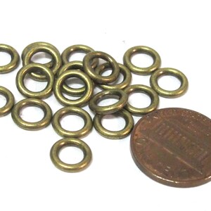 May include: A collection of small, brass-colored metal eyelets, scattered on a white surface. A copper penny is next to the eyelets for size comparison. The eyelets are circular with a hole in the center.