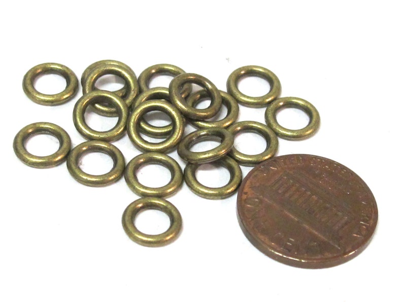 May include: A collection of small, brass-colored metal eyelets, scattered on a white surface. A copper penny is next to the eyelets for size comparison. The eyelets are circular with a hole in the center.