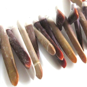 May include: Close-up of a necklace featuring elongated, natural-looking beads. The beads are in shades of brown, purple, and white, with a smooth, polished appearance. The necklace is set against a white background, highlighting the colors and textures.