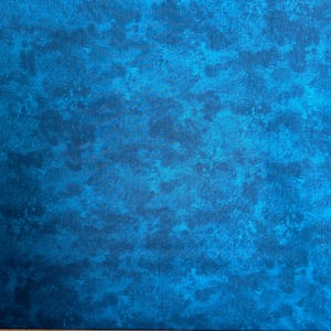 May include: A close-up of a blue fabric with a mottled pattern. The fabric has a mix of light and dark blue hues, creating a textured appearance. A wooden ruler with inch markings is at the bottom.