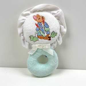 May include: A blue and white baby rattle with a Peter Rabbit embroidered design. The rattle has a white ruffle around the top and a white bow.