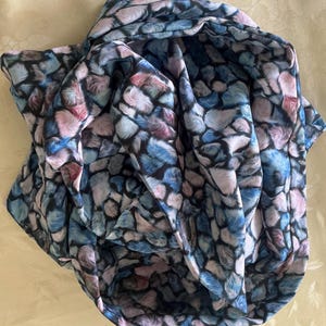 Könnte beinhalten: Ein drapierter Schal mit einem sich wiederholenden Muster aus blauen, rosa und lila Formen auf schwarzem Hintergrund. Der Schal besteht aus leichtem Stoff und hat ein weiches, strukturiertes Aussehen.
