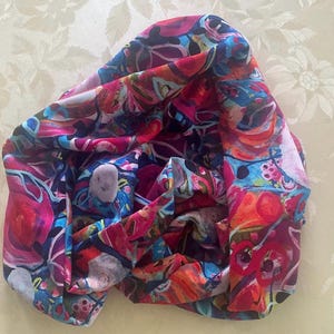 Könnte beinhalten: Ein farbenfrohes Tuch mit einem lebendigen Blumenmuster. Das Tuch zeigt eine Mischung aus roten, rosa, blauen und orangen Farbtönen, wodurch ein kühnes und künstlerisches Design entsteht. Der Stoff wirkt weich und fließend, ideal, um jedem Outfit einen Farbtupfer zu verleihen.
