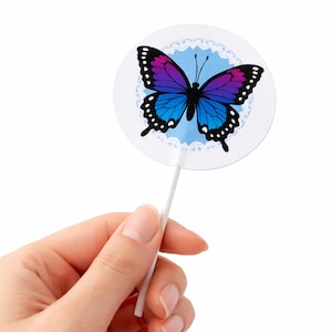 May include: A round, white butterfly-themed decoration on a stick. The butterfly has purple and blue wings with black detailing. The decoration is held by a hand against a white background.