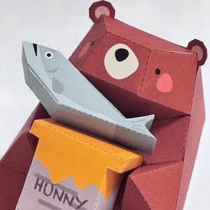 May include: A geometric papercraft bear in shades of red, holding a gray fish and a yellow honey pot. The honey pot has the word "HUNNY" printed on it. The bear has pink cheeks and small, rounded ears.