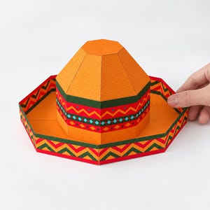Op de afbeelding: Een papieren sombrero-hoed met een feloranje basis en een geometrisch patroon in donkergroen, rood en lichtblauw. De hoed heeft een veelzijdige kroon en een brede rand. De tekst onderaan luidt: DIGITAL DOWNLOAD PDF FILE NO PHYSICAL ITEM.