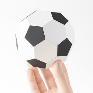 Könnte beinhalten: Ein Papierfußball mit schwarzen Fünfecken und weißen Sechsecken. Der Ball wird von einer Hand vor einem weißen Hintergrund gehalten. Der Ball hat eine geometrische Form und das Design ist ein klassisches Fußballmuster.