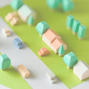 May include: A miniature papercraft village scene with houses, trees, and vehicles. The houses have peach-colored roofs and beige walls. Trees are green, and vehicles are blue and orange. The scene is set on a green and white paper background.